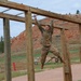 A soldier in military camouflage (OCP/MultiCam) uniform is traversing monkey bars on an outdoor obstacle course. The setting appears to be a military training facility with red rock terrain and pine trees visible in the background. The soldier is mid-traverse, suspended by both hands from the horizontal bars.