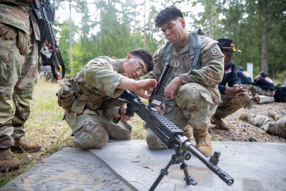 Two U.S. Army soldiers in MultiCam/OCP camouflage uniforms are crouched on a concrete pad, working together to assemble or inspect a light machine gun mounted on a bipod. The setting appears to be an outdoor training range surrounded by pine trees, with additional personnel visible in the background. This appears to be a weapons familiarization or qualification training exercise.