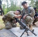 Two U.S. Army soldiers in MultiCam/OCP camouflage uniforms are crouched on a concrete pad, working together to assemble or inspect a light machine gun mounted on a bipod. The setting appears to be an outdoor training range surrounded by pine trees, with additional personnel visible in the background. This appears to be a weapons familiarization or qualification training exercise.