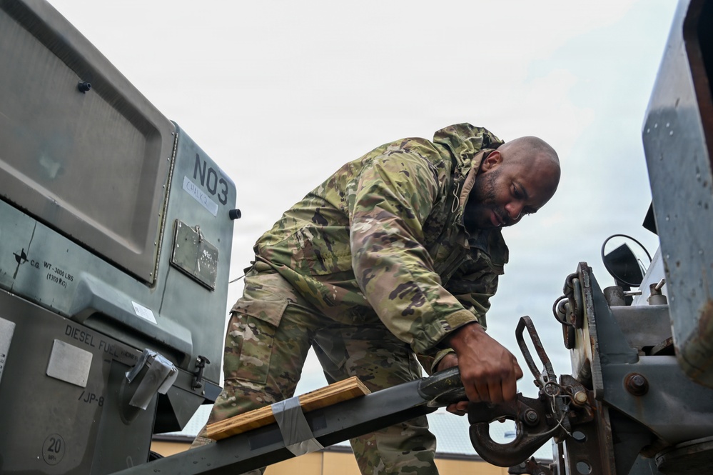 A military service member in OCP (Operational Camouflage Pattern) uniform is working on connecting or securing a tow hitch mechanism between two pieces of military equipment. The equipment visible includes what appears to be a military generator or power unit labeled 'N03' with markings indicating a weight of 3,000 lbs (1,360 kg) and 'DIESEL FUEL/JP-8'. The image is taken from a low angle, emphasizing the physical effort of the task.