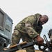 A military service member in OCP (Operational Camouflage Pattern) uniform is working on connecting or securing a tow hitch mechanism between two pieces of military equipment. The equipment visible includes what appears to be a military generator or power unit labeled 'N03' with markings indicating a weight of 3,000 lbs (1,360 kg) and 'DIESEL FUEL/JP-8'. The image is taken from a low angle, emphasizing the physical effort of the task.