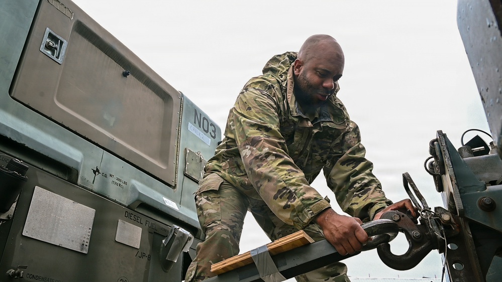 A military service member in MultiCam/OCP camouflage uniform is working with heavy equipment, manipulating a large hook or coupling mechanism on what appears to be military ground support equipment. The equipment is labeled 'N03' and includes markings indicating 'DIESEL FUEL' and weight specifications. The scene appears to be an outdoor military operations area under overcast skies.