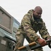 A military service member in MultiCam/OCP camouflage uniform is working with heavy equipment, manipulating a large hook or coupling mechanism on what appears to be military ground support equipment. The equipment is labeled 'N03' and includes markings indicating 'DIESEL FUEL' and weight specifications. The scene appears to be an outdoor military operations area under overcast skies.
