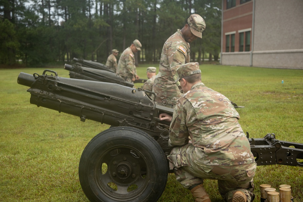 Several U.S. Army soldiers in MultiCam/OCP camouflage uniforms are working on a row of artillery pieces, likely M116 or similar howitzers, on a grassy area near a brick building. The soldiers appear to be conducting maintenance, inspection, or preparation of the cannons, possibly for a ceremonial salute. Spent brass casings are visible on the ground near the foreground cannon.