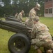 Several U.S. Army soldiers in MultiCam/OCP camouflage uniforms are working on a row of artillery pieces, likely M116 or similar howitzers, on a grassy area near a brick building. The soldiers appear to be conducting maintenance, inspection, or preparation of the cannons, possibly for a ceremonial salute. Spent brass casings are visible on the ground near the foreground cannon.