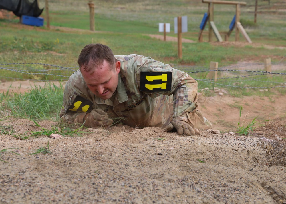 A soldier in military camouflage uniform is low-crawling under barbed wire on a sandy obstacle course. The individual is wearing number '11' markers in yellow on black, indicating a competition or training event. The setting appears to be a military training facility with various obstacles visible in the background.