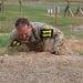 A soldier in military camouflage uniform is low-crawling under barbed wire on a sandy obstacle course. The individual is wearing number '11' markers in yellow on black, indicating a competition or training event. The setting appears to be a military training facility with various obstacles visible in the background.