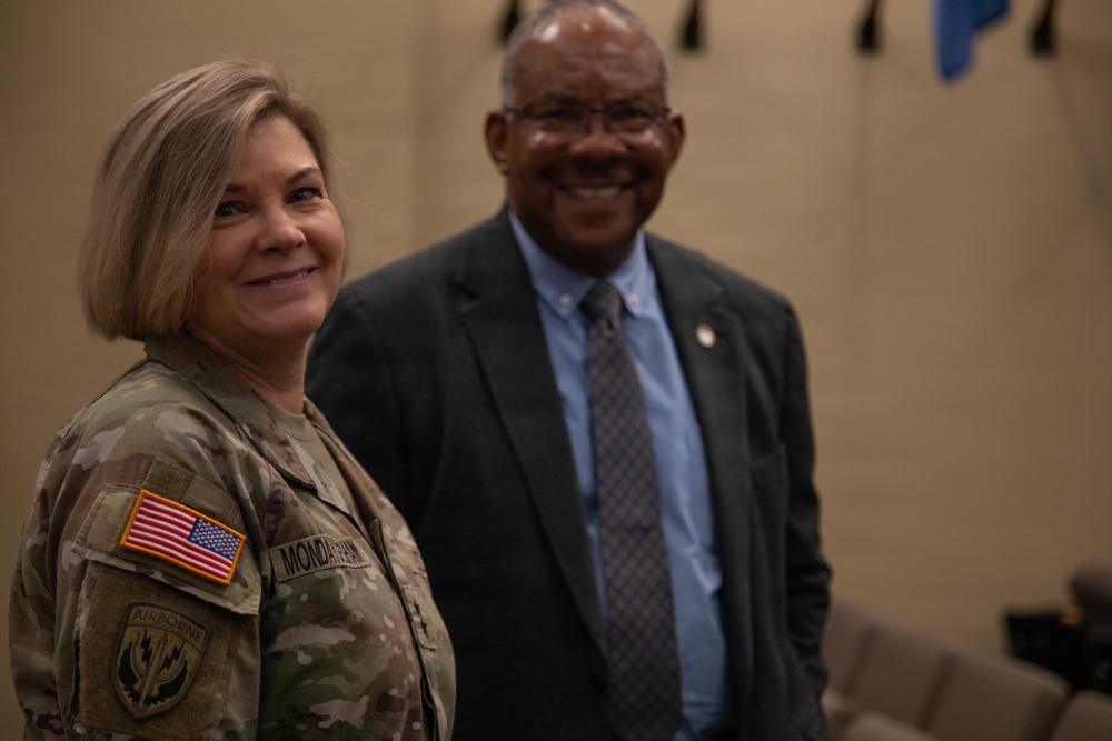 Two individuals are posing together in what appears to be a military or official setting. The woman on the left is wearing a U.S. Army multicam (OCP) uniform with an American flag patch, an Airborne tab, and a name tape reading 'MONDAYSMITH' or similar. The man on the right is dressed in civilian business attire consisting of a dark suit and tie.