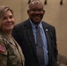 Two individuals are posing together in what appears to be a military or official setting. The woman on the left is wearing a U.S. Army multicam (OCP) uniform with an American flag patch, an Airborne tab, and a name tape reading 'MONDAYSMITH' or similar. The man on the right is dressed in civilian business attire consisting of a dark suit and tie.