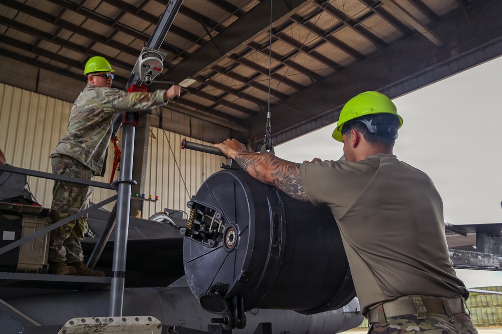Two military personnel wearing green hard hats and military uniforms are working together to install or remove a large cylindrical component, likely an aircraft gun system or sensor pod, using a hoist/crane mechanism inside a maintenance hangar. The worker in the foreground has visible tattoos on his arm and is steadying the component while the worker in the background operates the lifting equipment from an elevated platform. The setting appears to be a military aircraft maintenance facility.