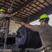 Two military personnel wearing green hard hats and military uniforms are working together to install or remove a large cylindrical component, likely an aircraft gun system or sensor pod, using a hoist/crane mechanism inside a maintenance hangar. The worker in the foreground has visible tattoos on his arm and is steadying the component while the worker in the background operates the lifting equipment from an elevated platform. The setting appears to be a military aircraft maintenance facility.