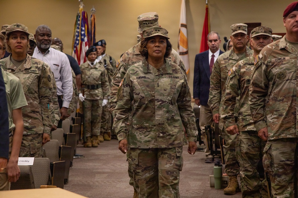 A formal U.S. Army military ceremony is taking place in an indoor venue, likely a conference or assembly hall. Soldiers in OCP (Operational Camouflage Pattern) uniforms are standing at attention or moving in formation, with a central female soldier bearing the name tape 'WALLACE' and a one-star general rank insignia prominently visible. Military flags and a color guard are visible in the background, indicating an official change of command or similar ceremonial event.