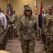 A formal U.S. Army military ceremony is taking place in an indoor venue, likely a conference or assembly hall. Soldiers in OCP (Operational Camouflage Pattern) uniforms are standing at attention or moving in formation, with a central female soldier bearing the name tape 'WALLACE' and a one-star general rank insignia prominently visible. Military flags and a color guard are visible in the background, indicating an official change of command or similar ceremonial event.