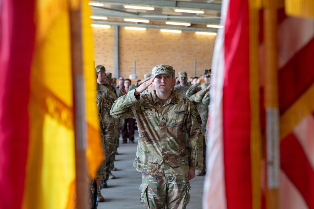 A U.S. Army soldier named Martin, wearing Operational Camouflage Pattern (OCP) uniform, renders a salute during a formal military ceremony. The soldier is framed between two flags — what appears to be a unit guidon and the U.S. flag — with rows of saluting soldiers visible in the background. The setting appears to be an indoor military facility or hangar with brick walls and fluorescent lighting.