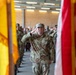A U.S. Army soldier named Martin, wearing Operational Camouflage Pattern (OCP) uniform, renders a salute during a formal military ceremony. The soldier is framed between two flags — what appears to be a unit guidon and the U.S. flag — with rows of saluting soldiers visible in the background. The setting appears to be an indoor military facility or hangar with brick walls and fluorescent lighting.