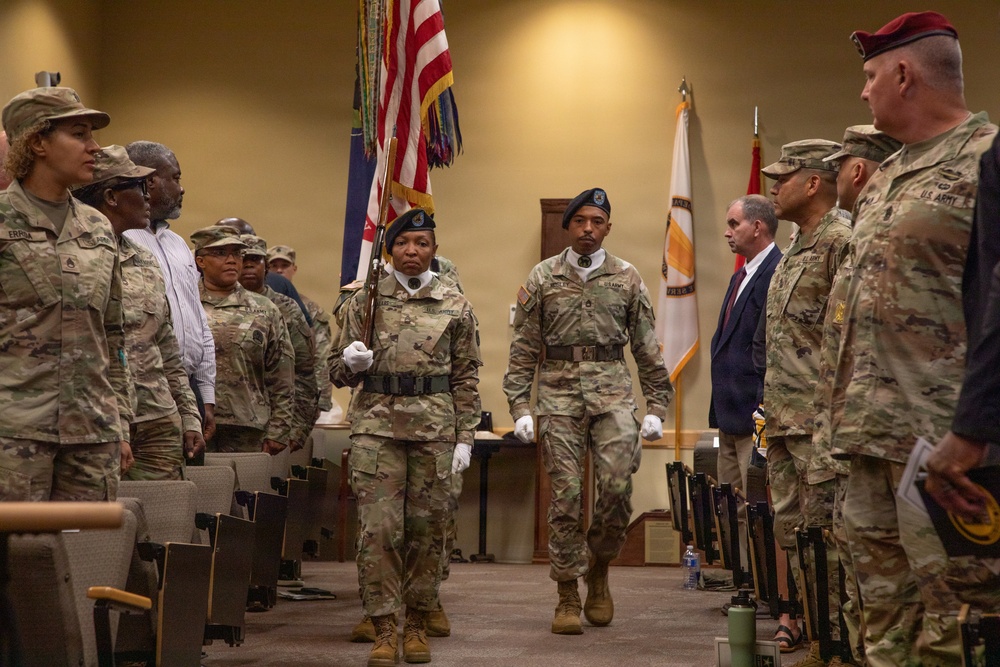 A U.S. Army military ceremony is taking place indoors, likely a change of command or formal military event. A color guard is marching down a center aisle carrying the American flag and other military colors, flanked by soldiers standing at attention. Both uniformed military personnel and civilian attendees are present in the room.