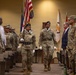 A U.S. Army military ceremony is taking place indoors, likely a change of command or formal military event. A color guard is marching down a center aisle carrying the American flag and other military colors, flanked by soldiers standing at attention. Both uniformed military personnel and civilian attendees are present in the room.