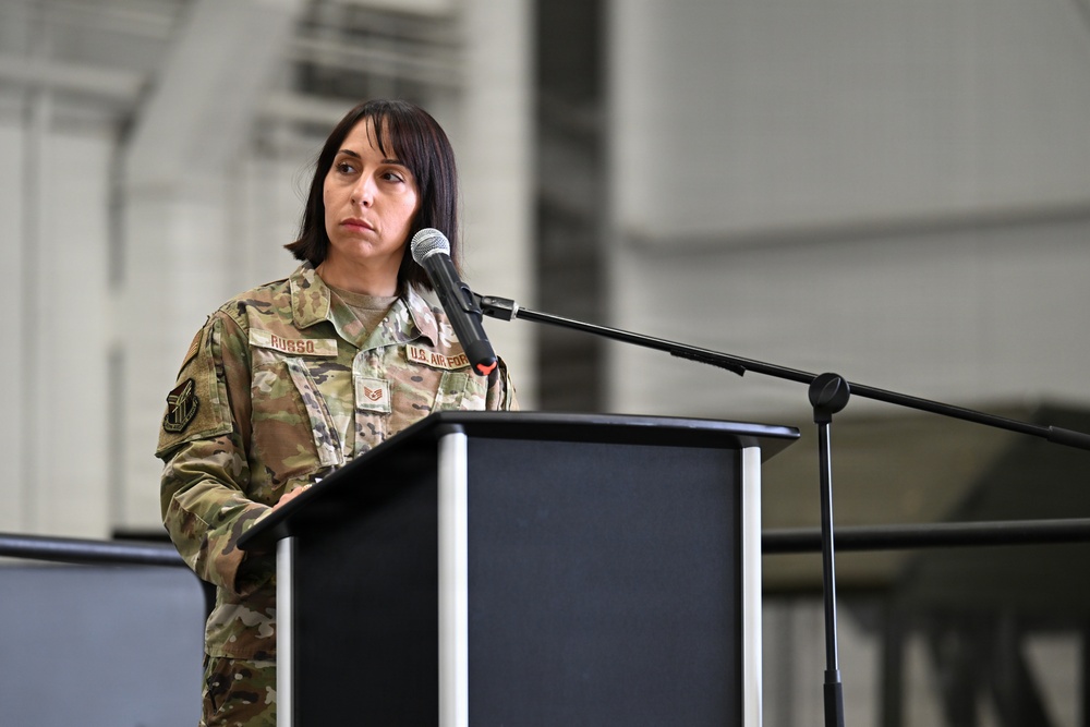 A U.S. Air Force service member stands at a black podium with a microphone, appearing to speak or prepare to speak at a formal military ceremony or event. The individual is wearing an OCP (Operational Camouflage Pattern) uniform with a name tape reading 'RUSSO' and a 'U.S. AIR FORCE' tape visible. The setting appears to be inside a large military hangar or similar facility.