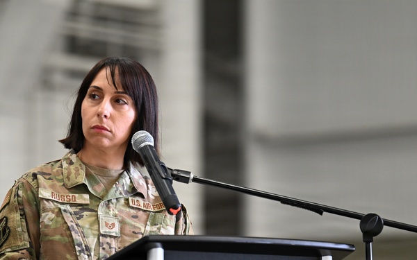 A U.S. Air Force service member stands at a black podium with a microphone, appearing to speak or prepare to speak at a formal military ceremony or event. The individual is wearing an OCP (Operational Camouflage Pattern) uniform with a name tape reading 'RUSSO' and a 'U.S. AIR FORCE' tape visible. The setting appears to be inside a large military hangar or similar facility.