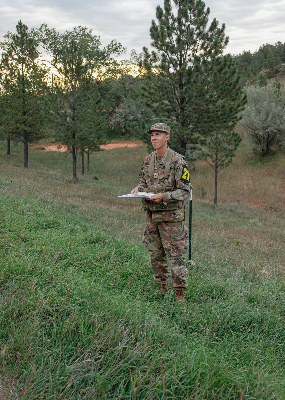 A soldier in U.S. Army OCP (Operational Camouflage Pattern) uniform stands in an open grassy field surrounded by pine trees at dusk or dawn. The individual is holding what appears to be a map or document and is standing next to a marker flag with the number '21' visible on their shoulder patch. This appears to be a land navigation training exercise.