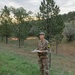 A soldier in U.S. Army OCP (Operational Camouflage Pattern) uniform stands in an open grassy field surrounded by pine trees at dusk or dawn. The individual is holding what appears to be a map or document and is standing next to a marker flag with the number '21' visible on their shoulder patch. This appears to be a land navigation training exercise.