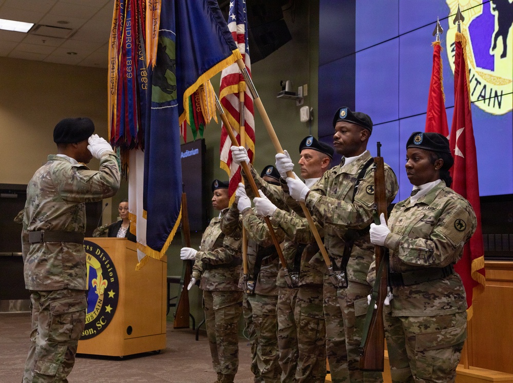 A U.S. Army color guard ceremony is taking place indoors at what appears to be an auditorium at Fort Jackson, South Carolina. Multiple soldiers in OCP (Operational Camouflage Pattern) uniforms and black berets are presenting colors while one soldier renders a salute. The ceremony appears to be an official military function, likely a change of command or activation ceremony.