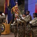 A U.S. Army color guard ceremony is taking place indoors at what appears to be an auditorium at Fort Jackson, South Carolina. Multiple soldiers in OCP (Operational Camouflage Pattern) uniforms and black berets are presenting colors while one soldier renders a salute. The ceremony appears to be an official military function, likely a change of command or activation ceremony.