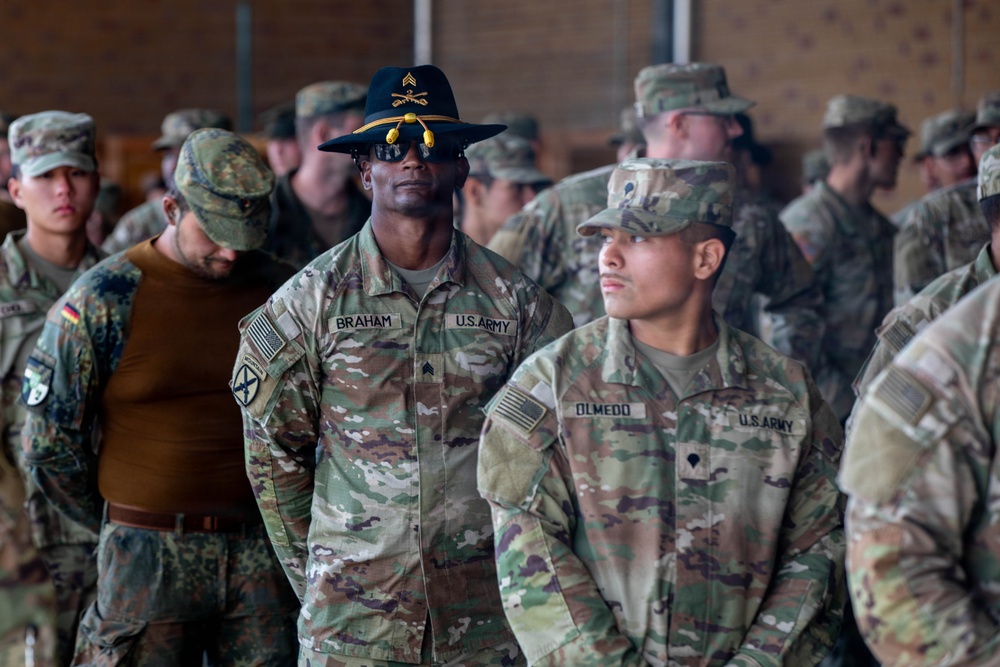 A group of U.S. Army soldiers and what appears to be at least one German Bundeswehr soldier are assembled in what looks like a military hangar or large indoor facility. Two U.S. Army soldiers are prominently visible in the foreground — one named BRAHAM wearing a distinctive cavalry Stetson hat with sergeant chevrons, and another named OLMEDO. The gathering appears to be a formal military formation or briefing event.