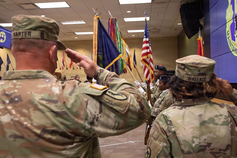 A U.S. Army military ceremony is taking place indoors, featuring a color guard presenting multiple flags including the American flag and unit guidons. Soldiers in Army Combat Uniforms (ACU/OCP pattern) are rendering salutes toward the color guard. Name tapes visible on personnel include 'HARTER' and 'WALLACE'.