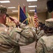 A U.S. Army military ceremony is taking place indoors, featuring a color guard presenting multiple flags including the American flag and unit guidons. Soldiers in Army Combat Uniforms (ACU/OCP pattern) are rendering salutes toward the color guard. Name tapes visible on personnel include 'HARTER' and 'WALLACE'.