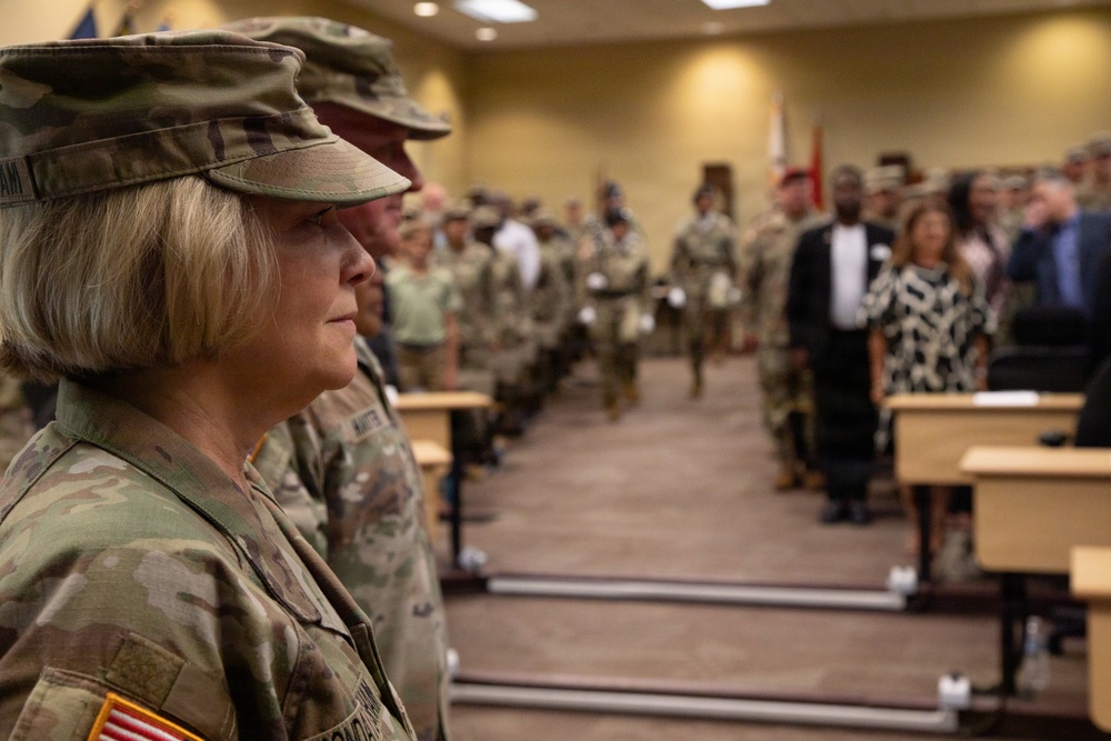 A military ceremony or formation is taking place inside what appears to be a conference or training room on a military installation. A female soldier in U.S. Army OCP (Operational Camouflage Pattern) uniform stands prominently in the foreground, with rows of uniformed soldiers and civilians visible in the background. The setting suggests an official event such as a promotion ceremony, change of command, or unit formation.