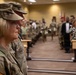 A military ceremony or formation is taking place inside what appears to be a conference or training room on a military installation. A female soldier in U.S. Army OCP (Operational Camouflage Pattern) uniform stands prominently in the foreground, with rows of uniformed soldiers and civilians visible in the background. The setting suggests an official event such as a promotion ceremony, change of command, or unit formation.