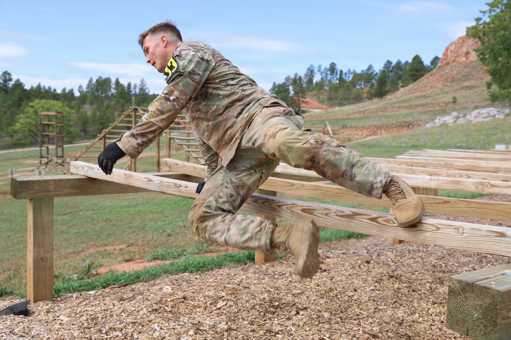 A soldier in camouflage uniform is navigating a military obstacle course, vaulting over a wooden beam barrier. The individual is captured mid-motion, leaning forward with hands on the beam and legs extended behind. The setting appears to be an outdoor military training facility with multiple obstacle structures visible in the background.