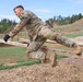 A soldier in camouflage uniform is navigating a military obstacle course, vaulting over a wooden beam barrier. The individual is captured mid-motion, leaning forward with hands on the beam and legs extended behind. The setting appears to be an outdoor military training facility with multiple obstacle structures visible in the background.