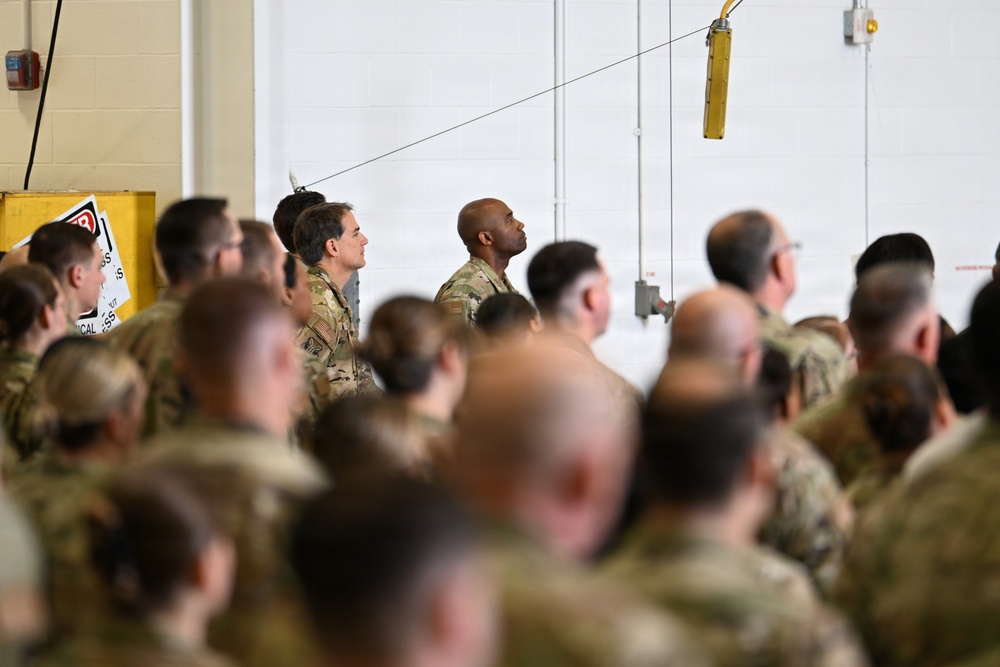 A large group of military personnel in camouflage uniforms are gathered inside what appears to be a hangar or large indoor facility. The crowd is densely packed, suggesting a formation, briefing, or ceremony. One soldier in the center-background is notably looking upward while others face forward.