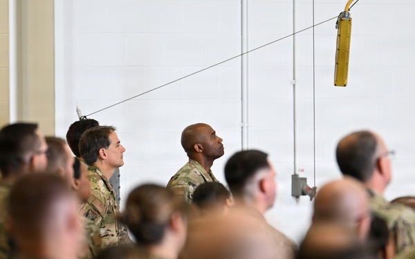 A large group of military personnel in camouflage uniforms are gathered inside what appears to be a hangar or large indoor facility. The crowd is densely packed, suggesting a formation, briefing, or ceremony. One soldier in the center-background is notably looking upward while others face forward.