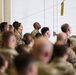 A large group of military personnel in camouflage uniforms are gathered inside what appears to be a hangar or large indoor facility. The crowd is densely packed, suggesting a formation, briefing, or ceremony. One soldier in the center-background is notably looking upward while others face forward.