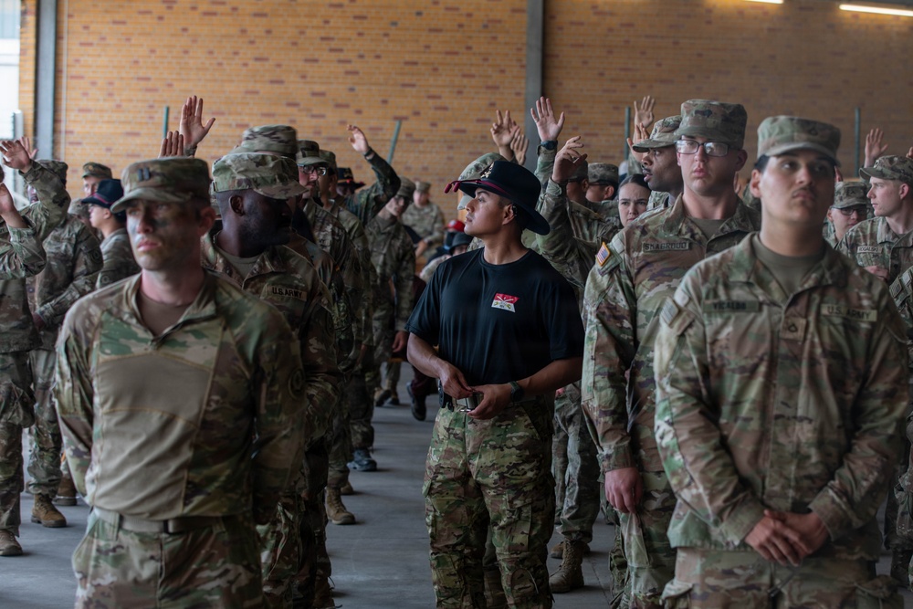 A large group of U.S. Army soldiers in Operational Camouflage Pattern (OCP) uniforms are assembled in what appears to be a covered outdoor formation area. One individual in the center stands out, wearing a black t-shirt, black campaign/drill sergeant hat, and OCP trousers, appearing to be an instructor or cadre member. Several soldiers in the background have their hands raised, suggesting a question-and-answer or accountability formation is taking place.