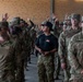A large group of U.S. Army soldiers in Operational Camouflage Pattern (OCP) uniforms are assembled in what appears to be a covered outdoor formation area. One individual in the center stands out, wearing a black t-shirt, black campaign/drill sergeant hat, and OCP trousers, appearing to be an instructor or cadre member. Several soldiers in the background have their hands raised, suggesting a question-and-answer or accountability formation is taking place.