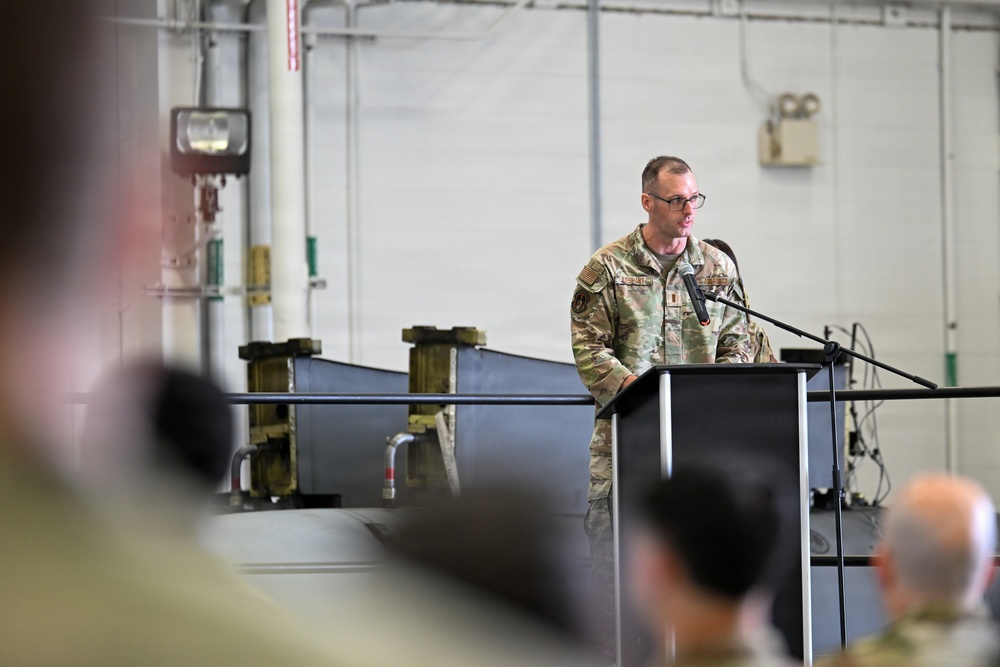 A military service member in OCP (Operational Camouflage Pattern) uniform stands at a podium with a microphone, appearing to deliver a speech or address. The setting is an industrial or hangar-type facility with a seated audience visible in the foreground. The event appears to be a formal military ceremony or briefing.