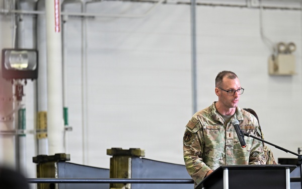 A military service member in OCP (Operational Camouflage Pattern) uniform stands at a podium with a microphone, appearing to deliver a speech or address. The setting is an industrial or hangar-type facility with a seated audience visible in the foreground. The event appears to be a formal military ceremony or briefing.
