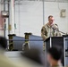 A military service member in OCP (Operational Camouflage Pattern) uniform stands at a podium with a microphone, appearing to deliver a speech or address. The setting is an industrial or hangar-type facility with a seated audience visible in the foreground. The event appears to be a formal military ceremony or briefing.