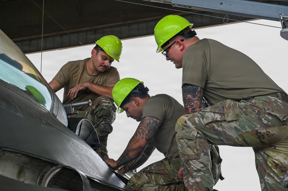 Three military personnel wearing bright green hard hats and camouflage/olive drab uniforms are performing maintenance work on a military aircraft, likely an F-16 Fighting Falcon. They are focused on a specific area of the aircraft's fuselage or wing root, with one individual using a tool. The work appears to be taking place under a hangar or shelter structure.