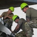 Three military personnel wearing bright green hard hats and camouflage/olive drab uniforms are performing maintenance work on a military aircraft, likely an F-16 Fighting Falcon. They are focused on a specific area of the aircraft's fuselage or wing root, with one individual using a tool. The work appears to be taking place under a hangar or shelter structure.