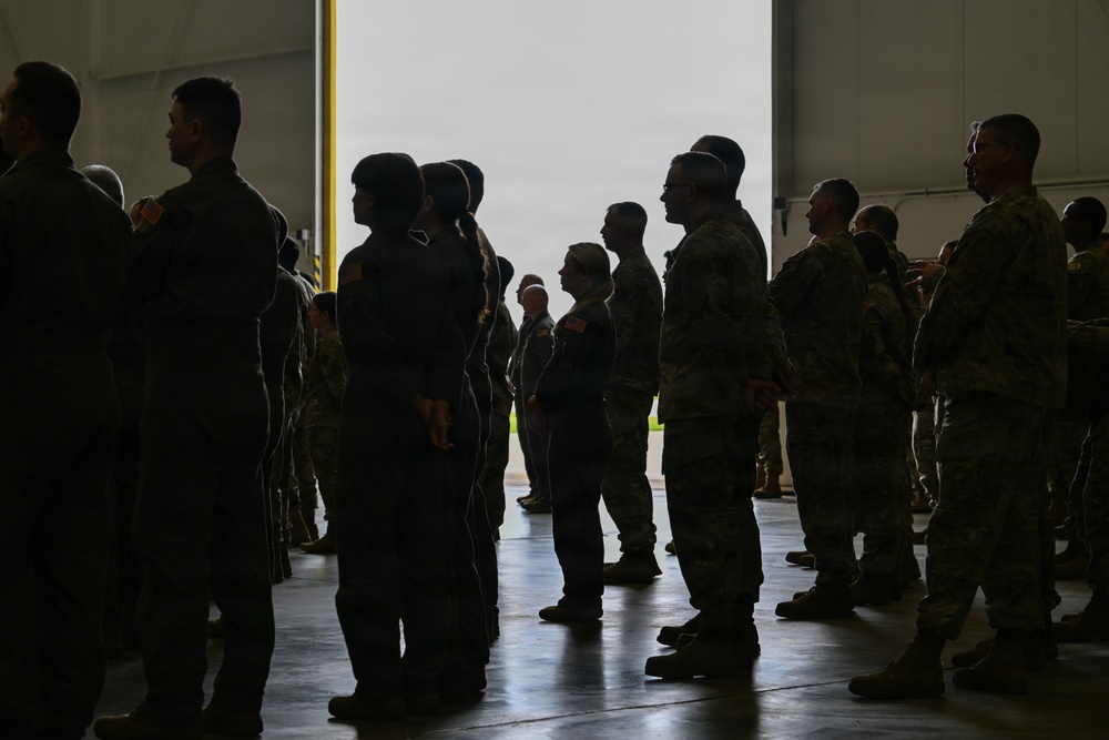 A group of military personnel in camouflage and flight suits are gathered inside what appears to be a large aircraft hangar. The personnel are standing in formation or loose groupings near a large open hangar door, which provides backlighting that partially silhouettes the figures. The setting suggests a briefing, formation, or pre-mission assembly.