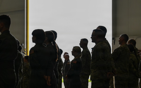 A group of military personnel in camouflage and flight suits are gathered inside what appears to be a large aircraft hangar. The personnel are standing in formation or loose groupings near a large open hangar door, which provides backlighting that partially silhouettes the figures. The setting suggests a briefing, formation, or pre-mission assembly.