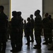 A group of military personnel in camouflage and flight suits are gathered inside what appears to be a large aircraft hangar. The personnel are standing in formation or loose groupings near a large open hangar door, which provides backlighting that partially silhouettes the figures. The setting suggests a briefing, formation, or pre-mission assembly.