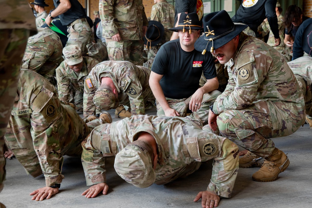 U.S. Army soldiers are engaged in what appears to be a combatives or physical training session inside a facility. Two individuals wearing distinctive black cavalry Stetson hats with crossed sabers insignia are supervising or instructing soldiers performing ground exercises. The scene depicts a structured military training environment with multiple participants.