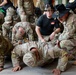 U.S. Army soldiers are engaged in what appears to be a combatives or physical training session inside a facility. Two individuals wearing distinctive black cavalry Stetson hats with crossed sabers insignia are supervising or instructing soldiers performing ground exercises. The scene depicts a structured military training environment with multiple participants.