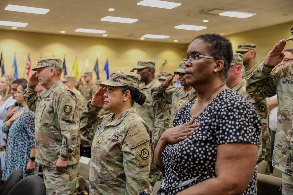 A formal military ceremony is taking place in an indoor venue, likely a conference or assembly room. Multiple U.S. Army soldiers in OCP (Operational Camouflage Pattern) uniforms are saluting while civilians stand with hands over their hearts, suggesting the playing of the National Anthem or Pledge of Allegiance. Various military unit flags are visible in the background.